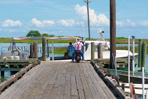 Tangier Island, Virginia / USA - June 21, 2020: A man on a small motorcycle talks with the captain of the “Miss Jenny” at the end of a wooden dock in this popular tourist destination in the Chesapeake Bay.