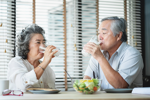 Smiling Asian senior man and woman sitting at table drinking glasses of milk and looking at each other. Cheerful Elderly couple enjoying healthy salad food while having breakfast at home.