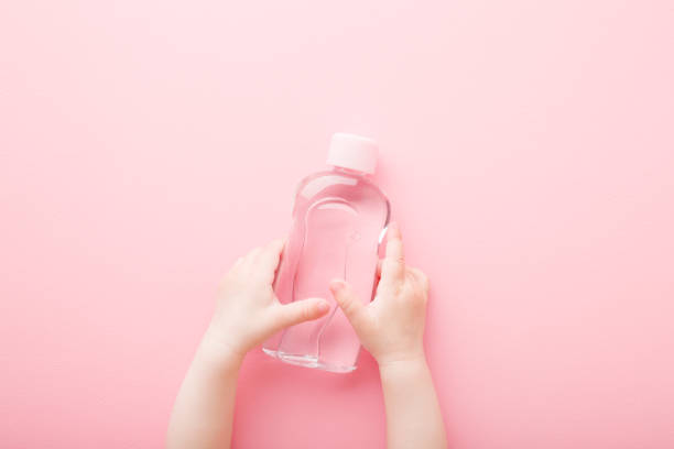 baby mädchen kleine hände halten transparente plastikflasche öl auf hellrosa tisch hintergrund. pastellfarbe. kümmern sie sich um weiche körperhaut. closeup. standpunkt geschossen. ansicht von oben nach unten. - massageöl stock-fotos und bilder