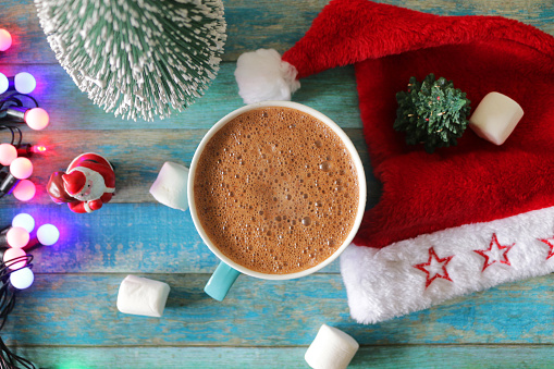 Stock photo showing an elevated view of a mug of hot cocoa drink beside a Santa hat, Christmas tree models, a Father Christmas figurine, string fairy light decoration surrounded by marshmallows, against a turquoise blue tongue and groove effect background.