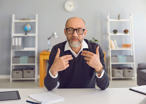 Mature man with a gray beard in spectacled looks at the camera, speaks with his partner sitting at a table in the office at home. An old male teacher communicates with gesticulating hands and gives a lesson to students.