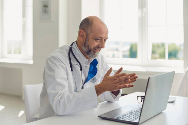 el médico de la tercera edad habla con el paciente usando la consulta de video webinar en línea portátil sentado en la oficina de la clínica. telemedicina. - patient medical exam meeting doctor fotografías e imágenes de stock