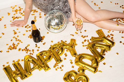 Hands and slim legs of young glamorous female with flute of champagne celebrating birthday while sitting on the floor with golden confetti