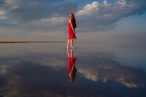 Elegant woman in silky red dress walking by a salt lake. Romantic mood. Water reflection of clouds and empty space. Holiday, vacation travel scene. lake Sivash