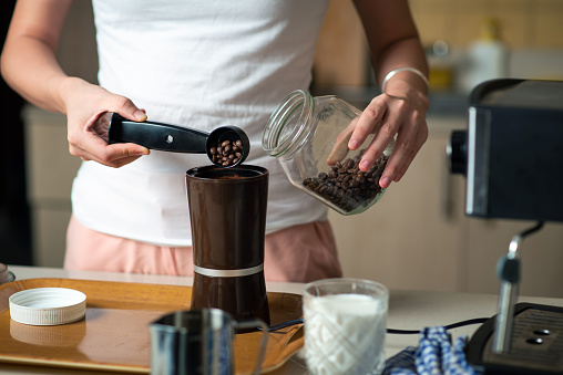 Woman inserting coffee beans into the grinder in the kitchen at home. Home barista and domestic lifestyle concept with unrecognizable person