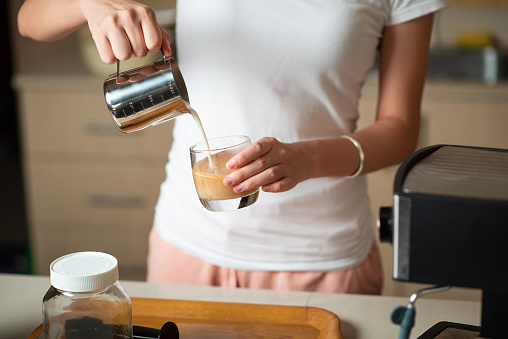 Woman pouring frothed milk into espresso while making a latte cappuccino coffee. Home barista and indoors lifestyle concept