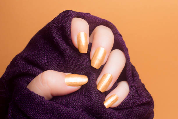 Female hand with beautiful gold colored nails in purple clothing on orange background, manicure concept.