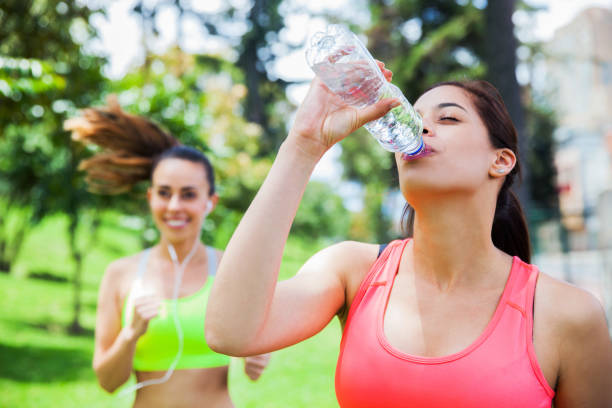 istock Young woman hydrating after jogging while another young athlete is running at background cheerfully 1255614086
