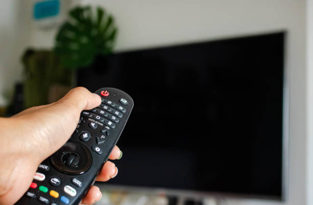 istock Watching Television Concept. Hand using a remote control in front of the TV screen. Relaxation in Modern Living Room. Focus on Remote. copy space. 1255614156