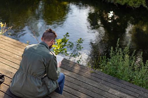 A man is reading a book by the water. He is wearing glasses and has a beard.