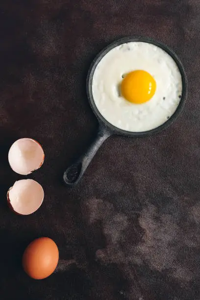 Photo of One fried egg in a small cast-iron pan on a dark rustic background. Top view, flat lay, copy space.