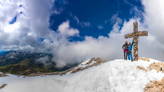 happy successful mountaineer hiking sporty fit people, couple cheering close to summit cross on mountain peak on sunny but cold and windy day in springtime, mountain range panorama alpe di siusi in background