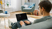Over the Shoulder Shot: Young Male Programmer Wearing Glasses Works on a Laptop Computer, Writing Scripts and Plug-ins. He's Sitting On a Couch in His Cozy Living Room at Home.