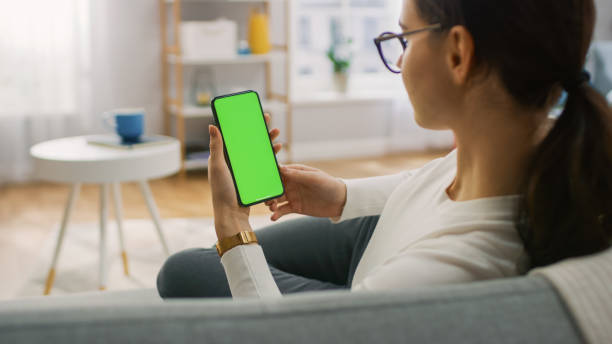 Young Woman at Home Uses Green Mock-up Screen Smartphone. She's Sitting On a Couch in His Cozy Living Room. Over the Shoulder Shot Young Woman at Home Uses Green Mock-up Screen Smartphone. She's Sitting On a Couch in His Cozy Living Room. Over the Shoulder Shot chroma key stock pictures, royalty-free photos & images