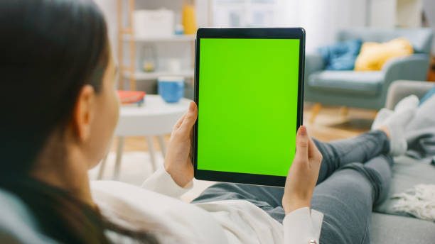istock Young Woman at Home Resting on a Couch Using with Green Mock-up Screen Tablet Computer in Vertical Portrait Mode. Woman Using Tablet Device, Browsing Internet, Watching Content, Videos. 1255633785