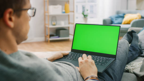 istock Man at Home Sitting on a Couch Working on Green Mock-up Screen Laptop Computer. Guy Using Laptop Device, Browsing Internet, Watching Content, Videos. Over the Shoulder Shot. 1255633786