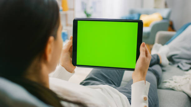 istock Young Woman at Home Resting on a Couch Using with Green Mock-up Screen Tablet Computer in Horizontal Landscape Mode. Woman Using Gestures with Touchscreen Device, Browsing Internet, Watching Content. 1255633823