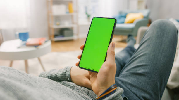 homme à la maison couché sur un divan utilisant le smartphone avec l’écran vert de maquette. au-dessus de la caméra de l’épaule shot. - caméra à lépaule photos et images de collection