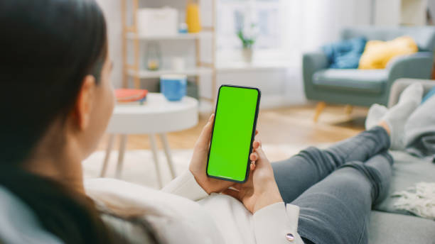 istock Young Woman at Home Resting on a Couch using with Green Mock-up Screen Smartphone. Girl Using Chroma Key Mobile Phone, Internet Browsing, Posting on Social Networks. Point of View Shot. 1255633841