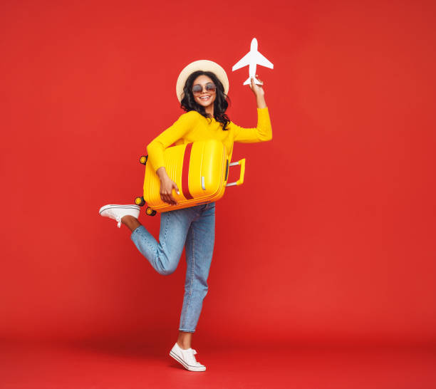 Ethnic traveler playing with toy plane Full body ethnic female in hat and sunglasses jumping   with suitcase and playing with toy airplane against red backdrop jump jet stock pictures, royalty-free photos & images