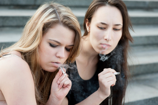 Two sad young girls smoke cigarettes outdoors