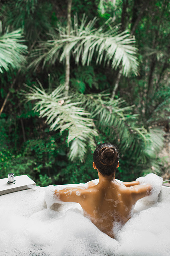 Woman relaxing in bath tub full of foam outdoors with jungle view. View from behind. Unrecognizable person. Beauty spa treatment, leisure time.