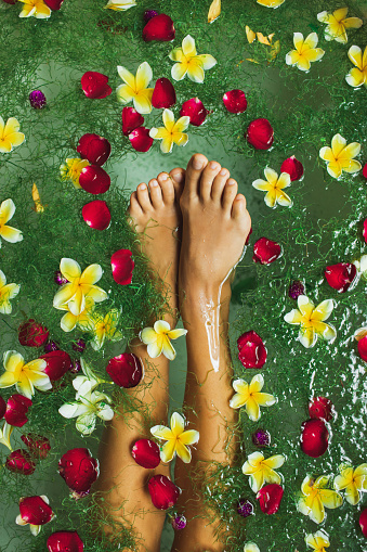 Woman legs in bath tub with tropical flowers, beauty spa treatment, pedicure, relaxation in hotel. Bali style. Rose red petals, yellow frangipani.