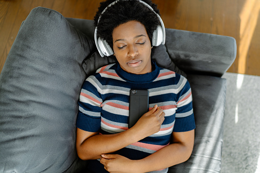 A Black young woman is lying on a sofa and resting while listening to music at home.