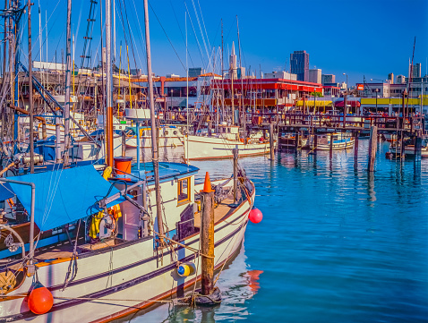 Fishing boats fill the marina at the famous Fisherman's Wharf at the waterfront of San Francisco, California. Tourists explore the stores and restaurants in front of the city skyline.