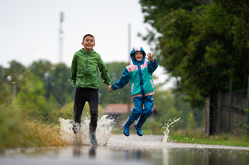 happy children siblings sister and brother jumping hand in hand through water puddle on rainy vacation day at home in rural landscape country road of small village