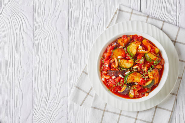 Stewed vegetables eggplant, tomatoes, zucchini with tomato sauce, garlic and herbs vegetable stew, eggplant, onion, zucchini with tomato sauce, garlic and herbs in a white bowl on a wooden table, landscape view from above, flat lay, free space ratatouille stock pictures, royalty-free photos & images