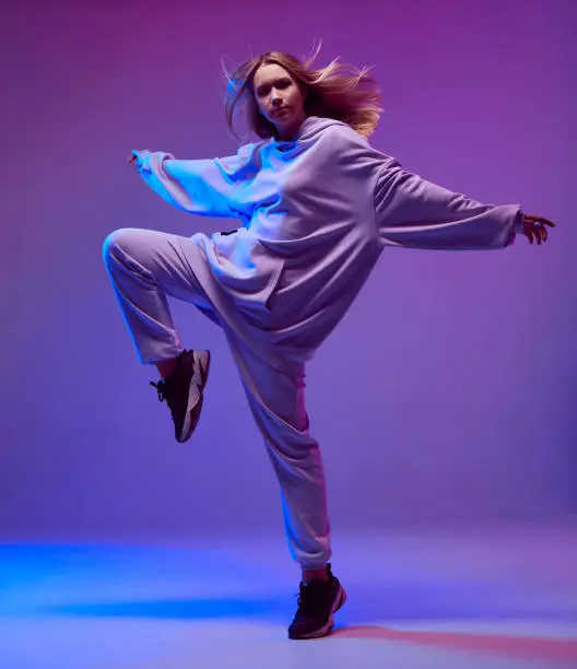 Photo of Portrait of a stylish young girl, cool dancing in a hoodie and with developing hair, on a neon background.
