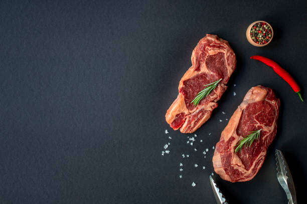 Two raw beef Rib eye steak with seasonings and tongs.