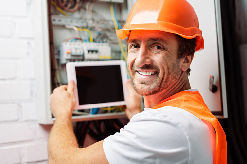 Selective focus of smiling electrician in hardhat holding digital tablet while working with electric panel