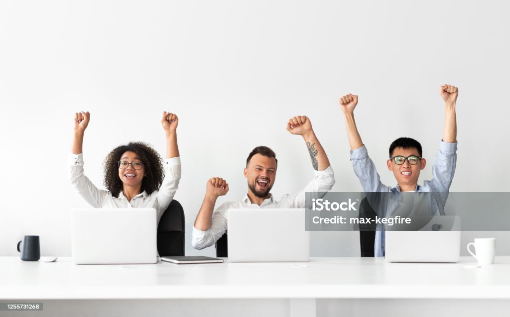 Happy colleagues sitting at laptops and celebrating success Happy excited modern business people with raised hands celebrating successful completion of project while sitting at table, with laptops in light office with white wall in background White Background Stock Photo