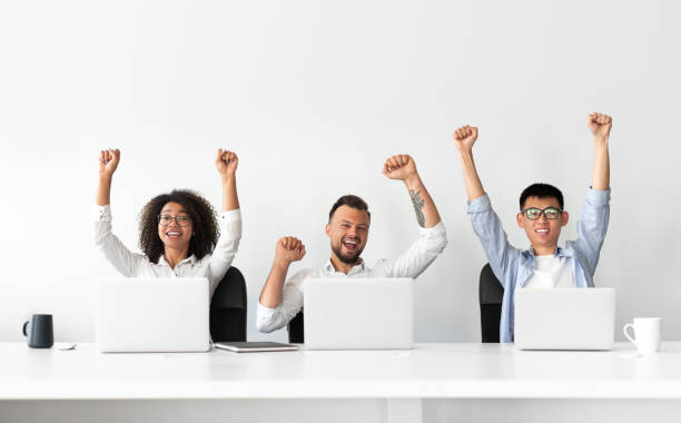 Happy colleagues sitting at laptops and celebrating success Happy excited modern business people with raised hands celebrating successful completion of project while sitting at table, with laptops in light office with white wall in background arms raised photos stock pictures, royalty-free photos & images