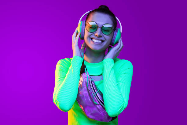 giovane ragazza positiva che indossa top e occhiali verde neon, godendo di ascoltare musica in cuffia con gli occhi chiusi, isolata su sfondo viola - purple belt foto e immagini stock