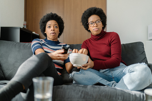 Black twin sisters are watching tv together at home.