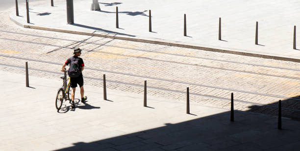 One young man walking next to his bike while crossing the street n a hot summer day , high angle rear view Belgrade, Serbia - July 9, 2020: One young man walking next to his bike while crossing the street with tram rail on a hot summer day , high angle rear view top view on people walking and cyclists riding stock pictures, royalty-free photos & images