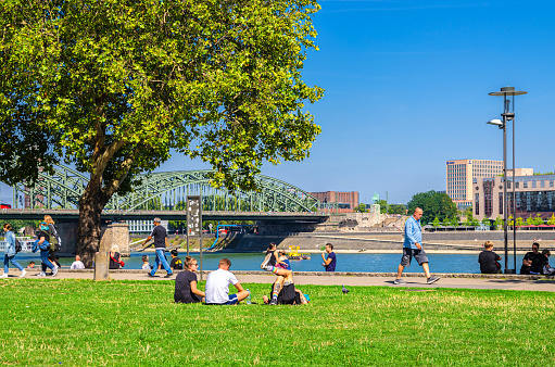 Cologne, Germany, August 23, 2019: people tourists walking down and having a rest on green grass lawn near Rhine river promenade in historical city centre, Hohenzollern Bridge and blue sky background