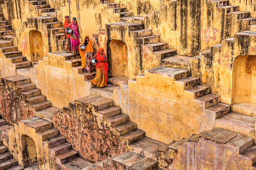 Indian women carrying water from stepwell near Jaipur, Rajasthan, India. Women and children often walk long distances to bring back jugs of water that they carry on their head. \nStepwells are wells in which the water may be reached by descending a set of steps.