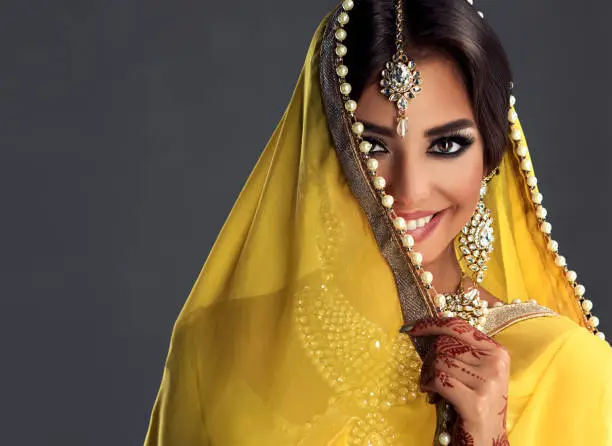 Black haired indian young woman dressed  in a posh yellow sari costume is shows toothy smile from behind traditional indian veil-dupatta. Sincere look. Indian tradition woman dress, henna-tattoo and stylish makeup.Asian beauty.