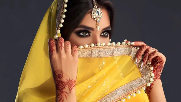 Black haired indian young woman dressed  in a posh yellow sari costume is hiding eyes behind traditional indian veil-dupatta. Indian tradition woman dress, henna-tattoo and stylish makeup.Asian beauty.