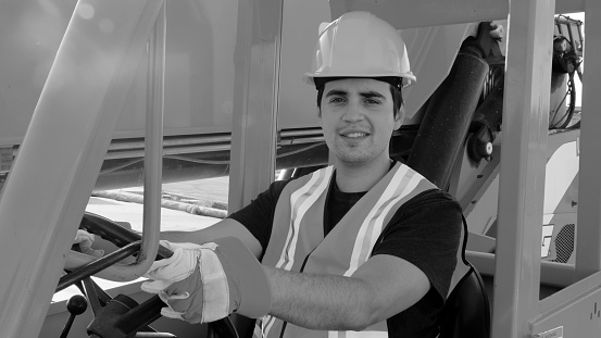 Black and white image of a Hispanic Young male construction worker looking at the camera smiling holding the  steering wheel of a big size forklift