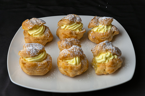 Completed golden brown homemade puff pastries are piped full with vanilla creme custard. The profiteroles are displayed on a white plate with a black background.