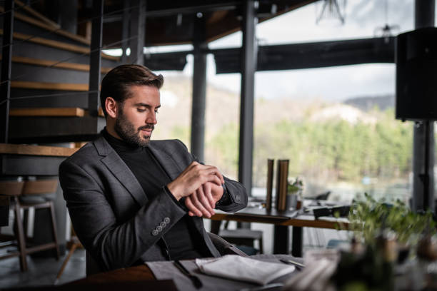 jeune homme d’affaires s’asseyant dans le restaurant et regardant le temps à la montre - tenue soignée photos et images de collection