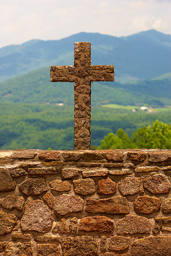 A stone cross on a stone wall in focus with a valley and mountain background.