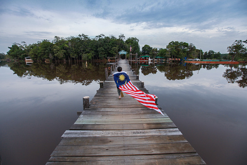 Malaysia's National Day and Malaysia Day is celebrated annually every 31st August and 16th September respectively. Malaysian flag 'Jalur Gemilang' can be found waving proudly not only in the cities but in the rural area as well. As seen here, a boy is running with a long Malaysian flag behind his back on a pier / jetty on a lake.