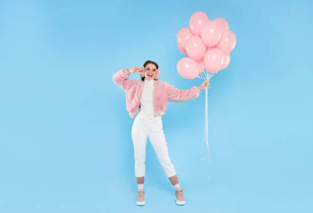 Photo of Full length portrait of young happy female wearing pink fluffy bomber and earwarmers, standing with pink birthday balloons, isolated on blue background