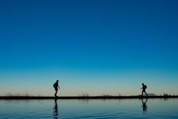 istock Silhouette Hikers walking towards one another on lake shore reflection with blue clear sky 1255782379